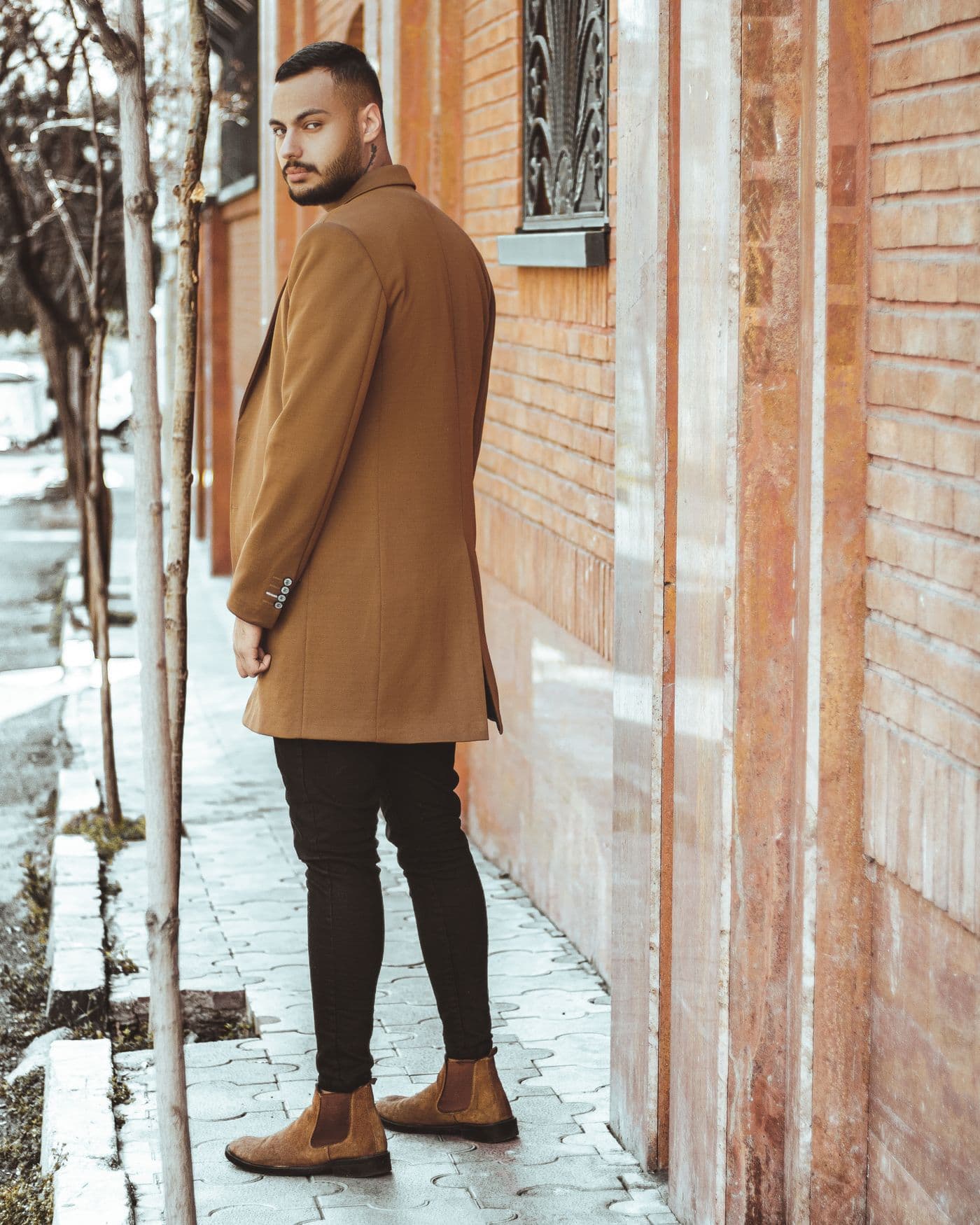 A man in a camel coat standing beside a brick building