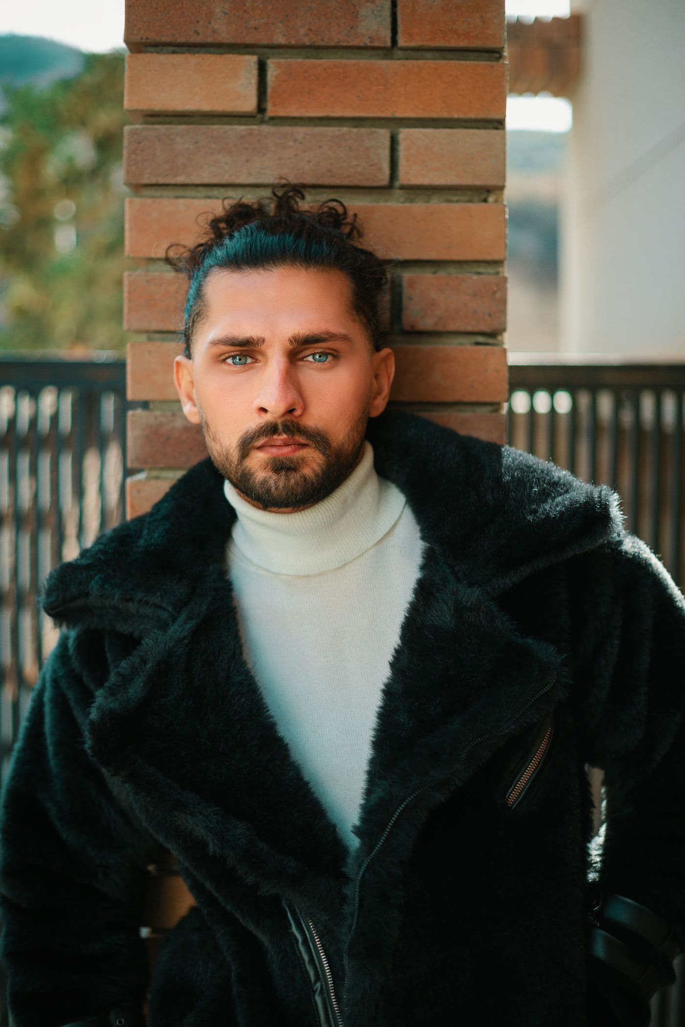 A man in a dark shearling coat over an ivory roll-neck against a brick wall