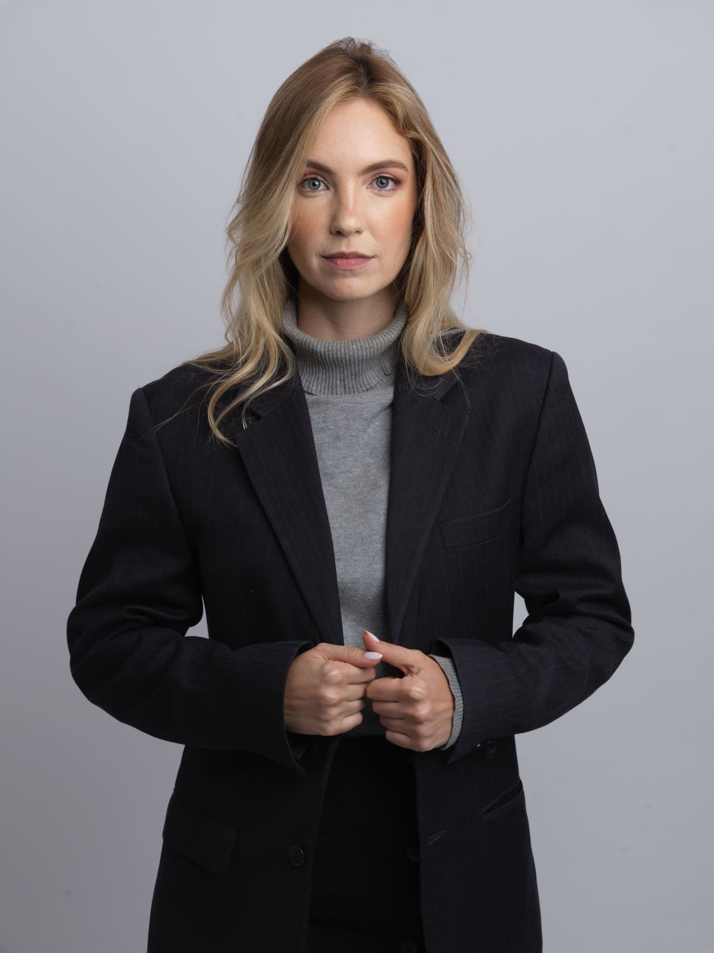A blonde woman in a black blazer over a grey roll-neck, studio portrait